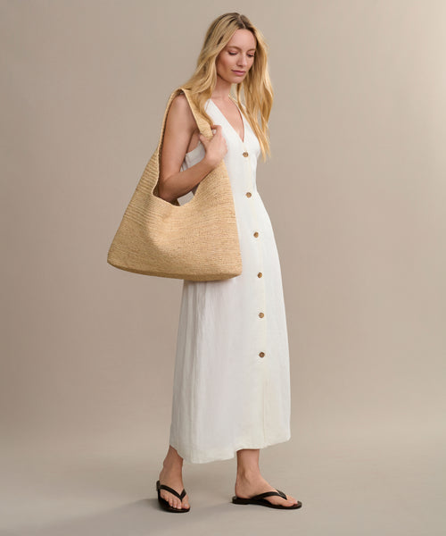 A woman carrying the Raffia Isla Bag over her shoulder, dressed in a sleeveless white button-front dress and black sandals, stands against a neutral background.