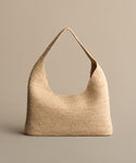 The Raffia Isla Bag, a natural woven piece with a wide single strap, is showcased upright against a simple beige background.