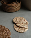 Three round Braided Raffia Coasters from Madagascar are stacked on a gray surface next to a larger basket, with another coaster partly visible in the foreground. Natural light creates soft shadows.