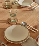 A wooden table set for two features white plates, cream-handled silverware, beige napkins, and glasses of water on Braided Raffia Coasters by Madagascar, organized neatly in natural light.