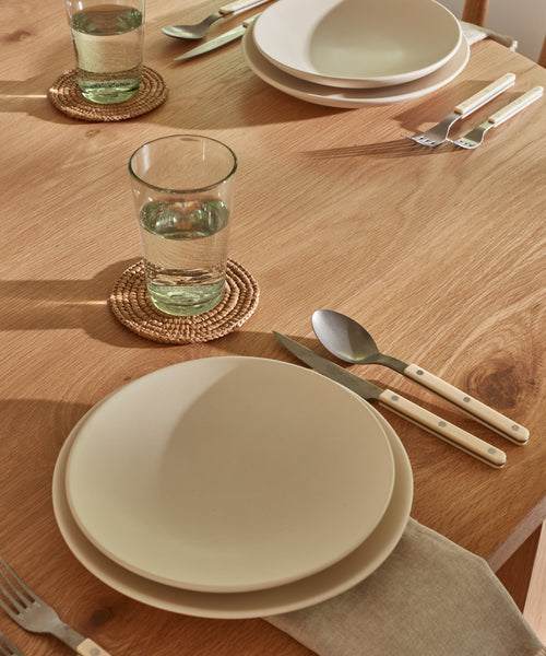 A wooden table set for two features white plates, cream-handled silverware, beige napkins, and glasses of water on Braided Raffia Coasters by Madagascar, organized neatly in natural light.