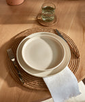 A glass of water sits on a Braided Raffia Coaster, with beige plates on a woven placemat, a knife and fork, and a white napkin—all arranged on a light wooden table.