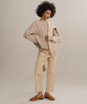 A woman with curly hair stands confidently against a plain light background, wearing the Ranger Trouser—a lightweight, fitted twill pant—paired with a chunky beige cable-knit sweater, matching sweater over her shoulder, and brown loafers.