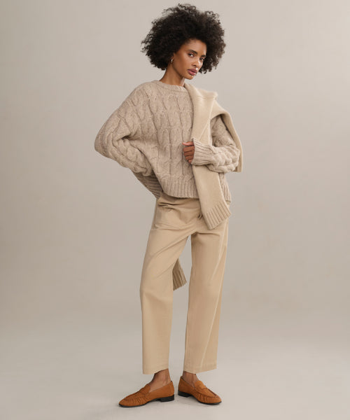 A woman with curly hair stands confidently against a plain light background, wearing the Ranger Trouser—a lightweight, fitted twill pant—paired with a chunky beige cable-knit sweater, matching sweater over her shoulder, and brown loafers.