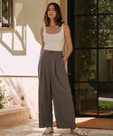 A woman poses casually outside a glass door, hands in pockets, wearing a white tank top and Relaxed Trouser with a high-waisted, wide-leg fit, paired with white sandals. Her shoulder-length hair frames her face.