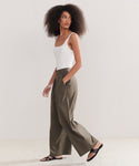 A woman with natural curly hair walks hands-in-pockets against a plain background, wearing a white sleeveless top and the Relaxed Trouser—high-waisted, raw silk wide-leg pants—paired with black sandals.
