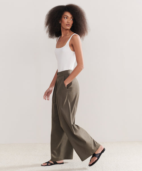 A woman with natural curly hair walks hands-in-pockets against a plain background, wearing a white sleeveless top and the Relaxed Trouser—high-waisted, raw silk wide-leg pants—paired with black sandals.