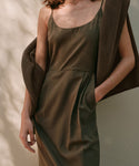 A woman wearing the Rove Dress, a sleeveless olive green raw silk dress, stands against a light wall with one hand in her pocket and a brown sweater over her shoulder.