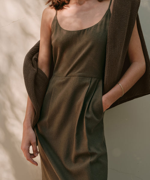 A woman wearing the Rove Dress, a sleeveless olive green raw silk dress, stands against a light wall with one hand in her pocket and a brown sweater over her shoulder.