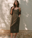 A woman stands outdoors in gentle sunlight, wearing the Rove Dress—an olive green sleeveless dress with pockets—in front of a pale wall with dappled shadows. A brown sweater is draped over her shoulders. A perfect spring-summer staple.