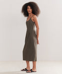 A woman with curly hair models the Rove Dress, a sleeveless olive green dress with thin straps, paired with black sandals—an easy spring-summer staple—while standing sideways against a plain light backdrop.