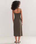 A woman with curly hair, facing away against a plain light background, wears the Rove Dress: a sleeveless olive green calf-length dress with thin straps and black sandals—a true spring-summer staple.