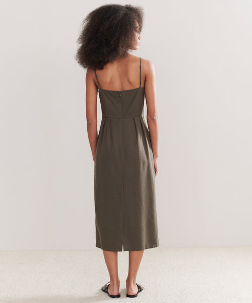 A woman with curly hair, facing away against a plain light background, wears the Rove Dress: a sleeveless olive green calf-length dress with thin straps and black sandals—a true spring-summer staple.