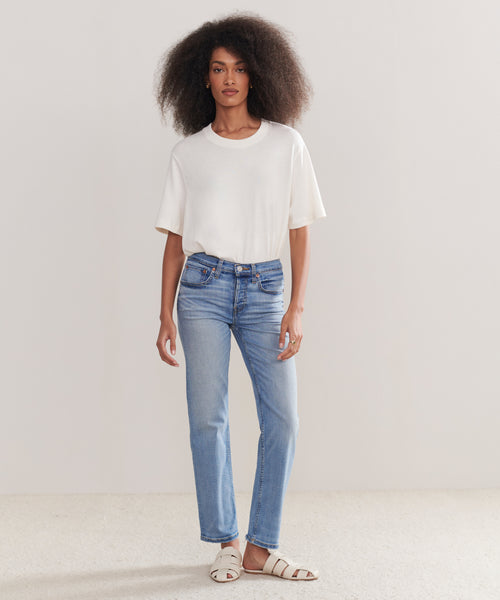 A woman with voluminous curly hair wears a plain white T-shirt, Mid Rise Stovepipe jeans, and white sandals while standing against a simple, light-colored background.