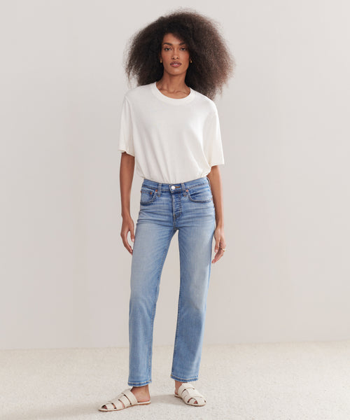 A woman with voluminous curly hair and a neutral expression stands against a plain light background, wearing a loose white t-shirt, Mid Rise Stovepipe jeans in a straight-leg fit, and white sandals.
