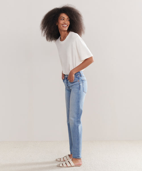A woman with natural curly hair, a white t-shirt, Mid Rise Stovepipe jeans, and white sandals stands smiling and looking sideways against a plain light background.