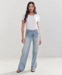 A woman with long, straight dark hair and a neutral expression stands against a plain white background, wearing a white short-sleeved t-shirt, light blue Mid Rise Wide Leg jeans, and black sandals.