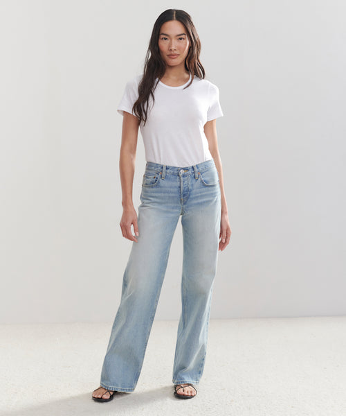 A woman with long, straight dark hair and a neutral expression stands against a plain white background, wearing a white short-sleeved t-shirt, light blue Mid Rise Wide Leg jeans, and black sandals.