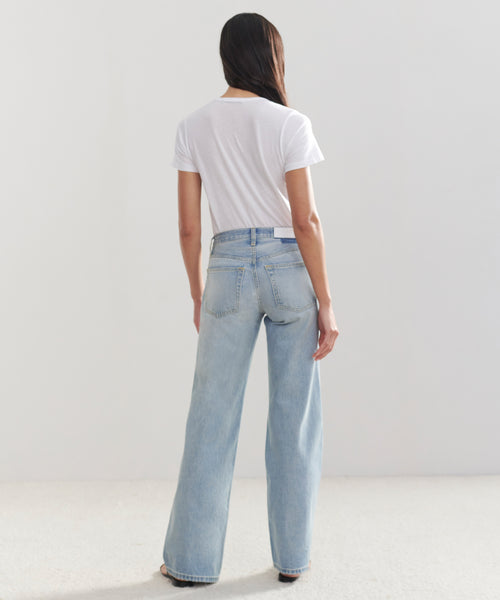 A person with long dark hair, wearing a white t-shirt and light blue Mid Rise Wide Leg jeans, stands facing away in a minimal, light-colored room.