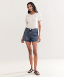 A woman with long dark hair stands in a minimal room wearing a plain white t-shirt, Mid Rise Boy Short dark denim shorts, and black sandals. She looks relaxed with one hand by her side and the other loosely bent.
