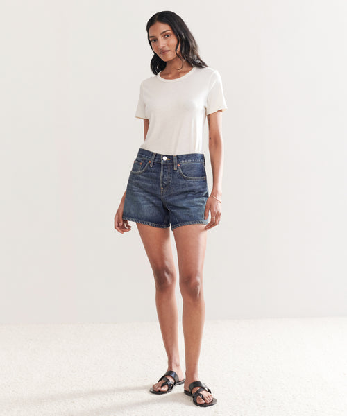 A woman with long dark hair stands in a minimal room wearing a plain white t-shirt, Mid Rise Boy Short dark denim shorts, and black sandals. She looks relaxed with one hand by her side and the other loosely bent.