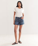 A woman with long dark hair and a confident look stands in a minimal, well-lit room wearing Mid Rise Boy Short Re/Done jeans made from organic cotton denim, a short-sleeve white t-shirt, and black sandals.