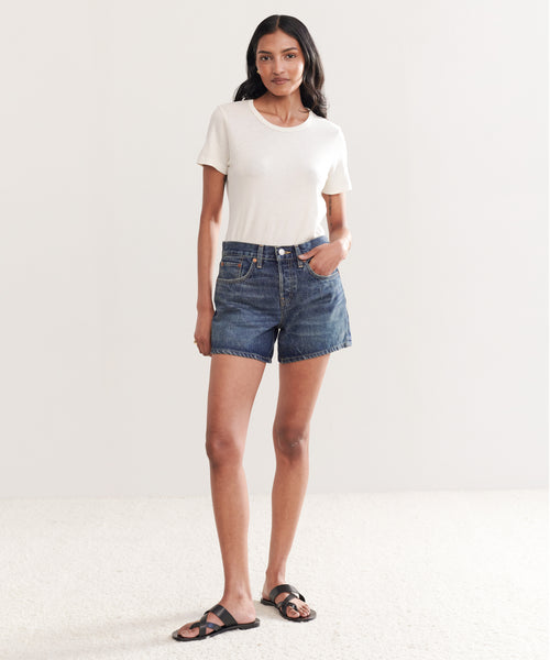 A woman with long dark hair and a confident look stands in a minimal, well-lit room wearing Mid Rise Boy Short Re/Done jeans made from organic cotton denim, a short-sleeve white t-shirt, and black sandals.