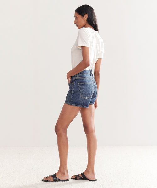 A person stands sideways on a light carpeted floor against a white wall, wearing a white t-shirt, the Mid Rise Boy Short in organic cotton denim, and black sandals, with one hand in their pocket.