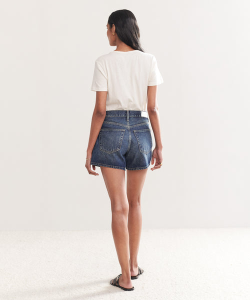A woman with long dark hair stands on a light-colored carpet, facing away, wearing a white t-shirt and Mid Rise Boy Shorts. She pairs the look with sandals against a plain white background.