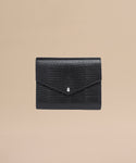 The Reece Leather Wallet, a black textured clutch with a flap closure and silver teardrop accent, is shown on a plain beige background—ideal as a mid-size wallet.