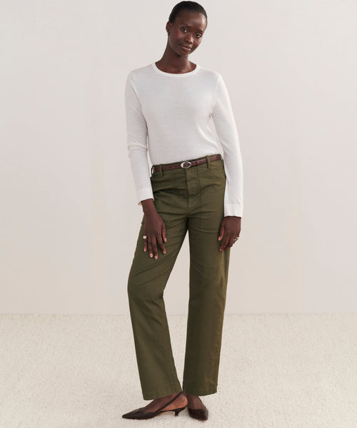 A woman stands against a light background in a white long-sleeve shirt, Relaxed Army Pant with a brown belt, and brown pointed-toe flats. She smiles softly with one hand resting on her leg.