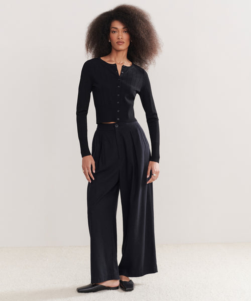 A woman with voluminous curly hair wears a fitted black button-up top and high-waisted Relaxed Trouser, paired with black flats, standing against a plain light background.