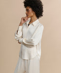 A woman with curly hair stands in profile against a beige background, wearing the loose-fitting, long-sleeve Remy Shirt with puffed sleeves and matching wide-leg pants. She rests one hand on her chin and appears thoughtful.