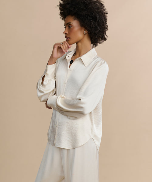 A woman with curly hair stands in profile against a beige background, wearing the loose-fitting, long-sleeve Remy Shirt with puffed sleeves and matching wide-leg pants. She rests one hand on her chin and appears thoughtful.