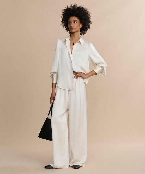 A woman with short curly hair poses against a beige background, wearing the Remy Shirt and wide-leg pants. She carries a black tote bag and wears black flats.
