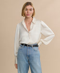 A woman with a short blonde bob wears the cream-colored satin Remy Shirt with puffed sleeves, tucked into high-waisted blue jeans and finished with a black belt, standing against a beige background with a neutral expression.