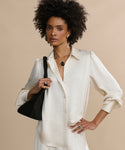 A woman with natural curly hair models the cream-colored Remy Shirt with puffed sleeves, white pants, and a black shoulder bag. She accessorizes with rings and a black pendant necklace, standing against a beige background.