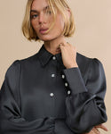 A woman with a short blonde bob models the Remy Shirt, a vintage-inspired satin top with puffed sleeves and pearl buttons. She stands against a beige background, touching her collar and gazing off to the side.