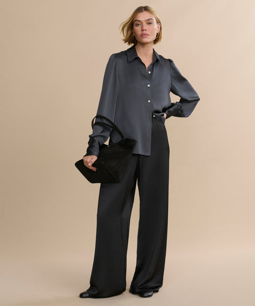 A woman with short blonde hair and a confident look stands against a beige background wearing the vintage-inspired satin Remy Shirt with puffed sleeves, wide-leg black pants, and holding a black handbag.