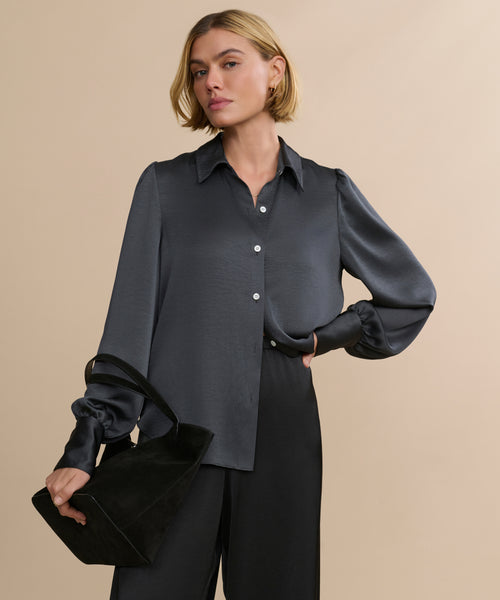 A woman with short blonde hair confidently models the vintage-inspired satin Remy Shirt with puffed sleeves and black pants, holding a black handbag against a plain beige background.