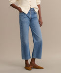 A person stands with hands in pockets, wearing the Ren jean in light blue—a high-rise, straight-leg style—paired with a cream button-up sweater and brown pointed-toe flats against a plain beige background.