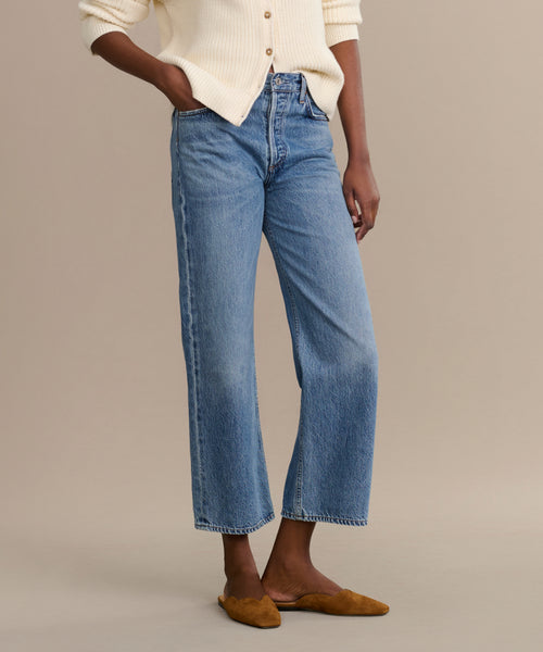 A person stands with hands in pockets, wearing the Ren jean in light blue—a high-rise, straight-leg style—paired with a cream button-up sweater and brown pointed-toe flats against a plain beige background.