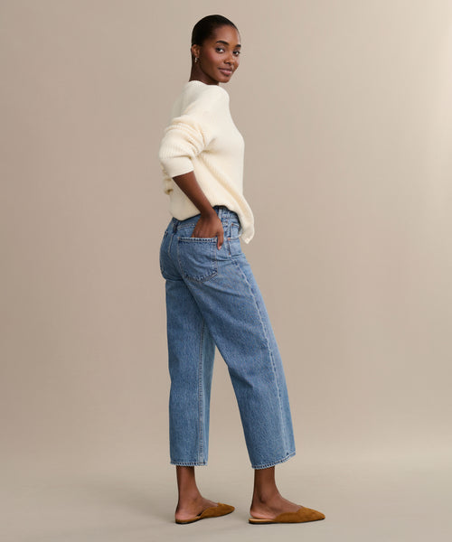 A woman stands sideways against a plain beige background, looking at the camera. She wears light blue Ren jeans with a wide-leg silhouette, a cream sweater, and brown flat shoes. Her hair is pulled back.