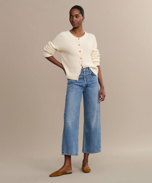 A person stands against a beige background wearing a cream button-up cardigan, brown flats, and the Ren jean—high-rise wide-leg jeans made from regenerative cotton—with one hand in their pocket and a neutral expression.