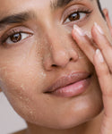 A close-up of a person gently touching their cheek, with smooth lips and brown eyes, showcasing Renew Face Polish featuring upcycled pineapple exfoliants applied to their skin.