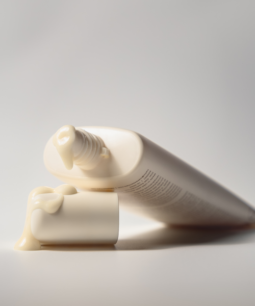 A white tube of Renew Face Polish lies on its side with the cap off, a small amount of pale cream—made with upcycled pineapple exfoliants—oozing onto the cap against a plain, light background.