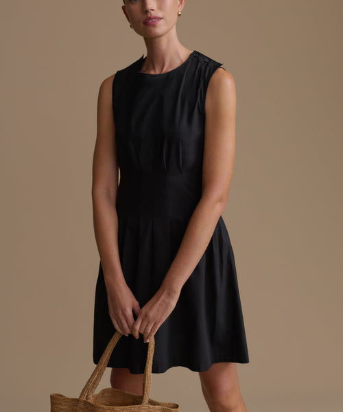 Woman wearing a black sleeveless dress holding a woven bag against a brown background