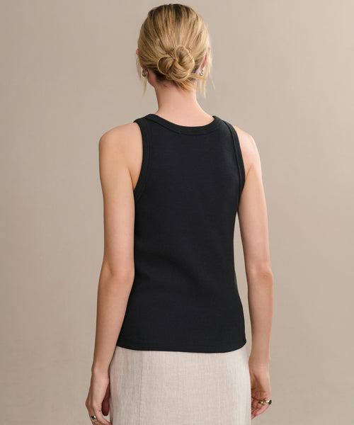 A woman with blonde hair in a low bun is seen from behind, wearing a sleeveless black Cotton Rib Tank and a light beige skirt, standing against a plain neutral background.
