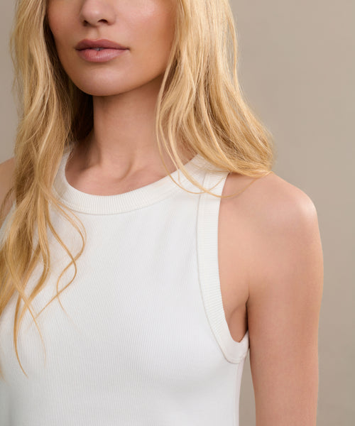 A woman with long blonde hair models the Cotton Rib Tank, a sleeveless white top, standing against a neutral beige background. Her face is partially visible with natural makeup and a relaxed expression.