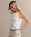 A woman with long blonde hair wears a sleeveless white Cotton Rib Tank and light beige belted pants, standing against a neutral background and looking down with her hands in her pockets.